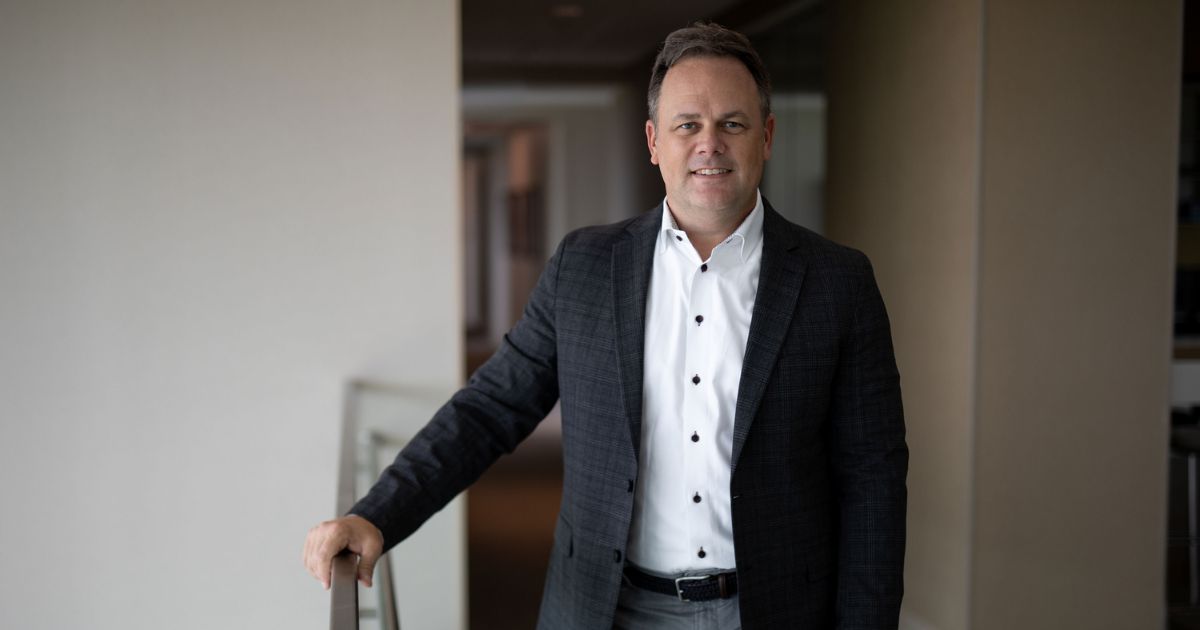Bryan Harris, CTO do SAS, posa sorridente em um ambiente corporativo moderno. Ele veste paletó xadrez escuro e camisa branca com botões pretos, apoiando uma das mãos no corrimão. Foto: Divulgação.
