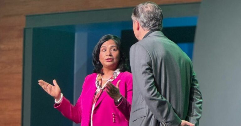 Ritu Jyoti, vice-presidente e gerente-geral da IDC, conversa no palco com Mike Capone, CEO da Qlik. Ela veste um blazer rosa vibrante e faz gestos com as mãos enquanto fala, enquanto Capone, de costas para a câmera, usa um terno cinza. Ambos estão em um ambiente de evento corporativo ou conferência (empresas)