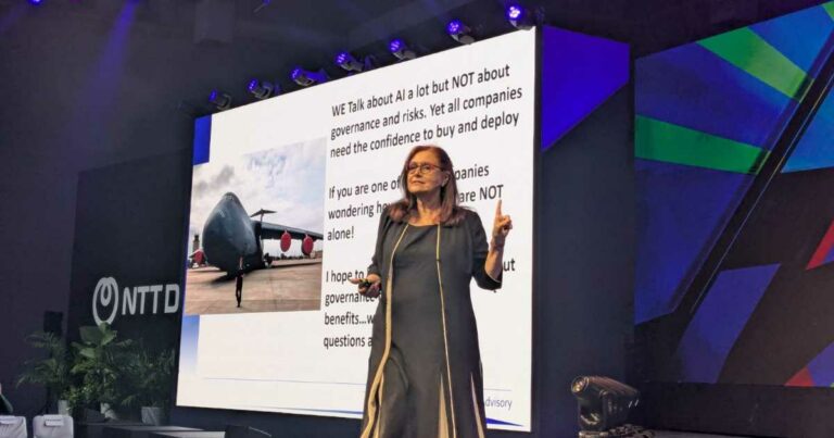 Kay Firth-Butterfield, CEO da Good Tech Advisory, está em pé no palco durante uma apresentação. Ela fala diante de um grande telão que exibe uma imagem de um avião militar e um texto sobre inteligência artificial, governança e riscos. O evento ocorre em um espaço moderno com iluminação de palco e o logotipo da NTT Data visível ao fundo (governança)