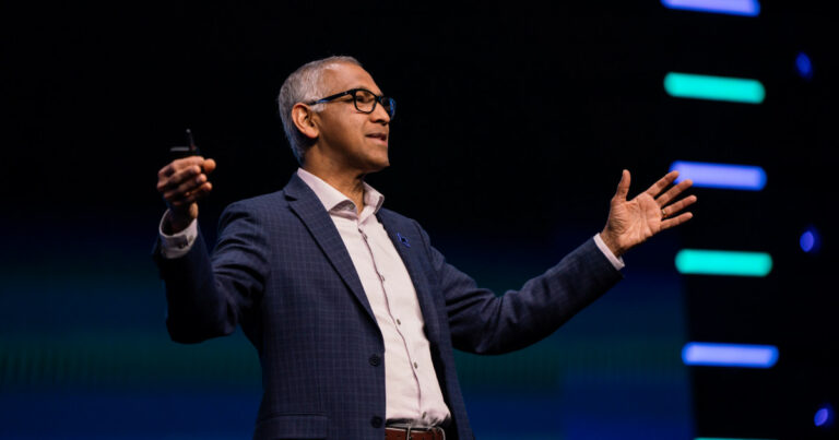 Rajiv Ramaswami, CEO da Nutanix, aparece em um palco durante uma apresentação. Ele está vestindo terno azul xadrez, camisa social branca e óculos, com os braços abertos em gesto expressivo, segurando um controle remoto em uma das mãos. Ao fundo, o cenário é escuro com luzes de LED coloridas em listras horizontais.