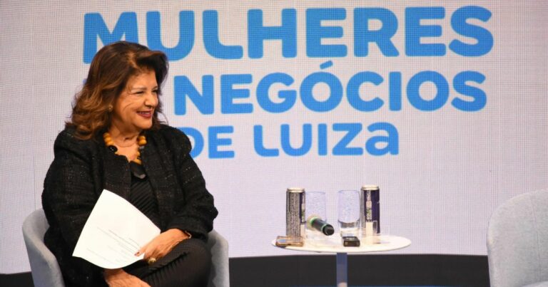 Mulher sentada numa cadeira, sorridente, segurando folhas de papel nas mãos. Ela usa um conjunto preto com colar de contas grandes em tons de amarelo e preto. Ao fundo, há um painel com o texto “Comunidade Seller Mulheres de Negócios de Luiza” em letras azuis. Ao lado dela, há uma mesa branca com duas latas, dois copos, um microfone e outros objetos pequenos. A cena decorre num ambiente de evento ou painel de discussão. (Magalu)