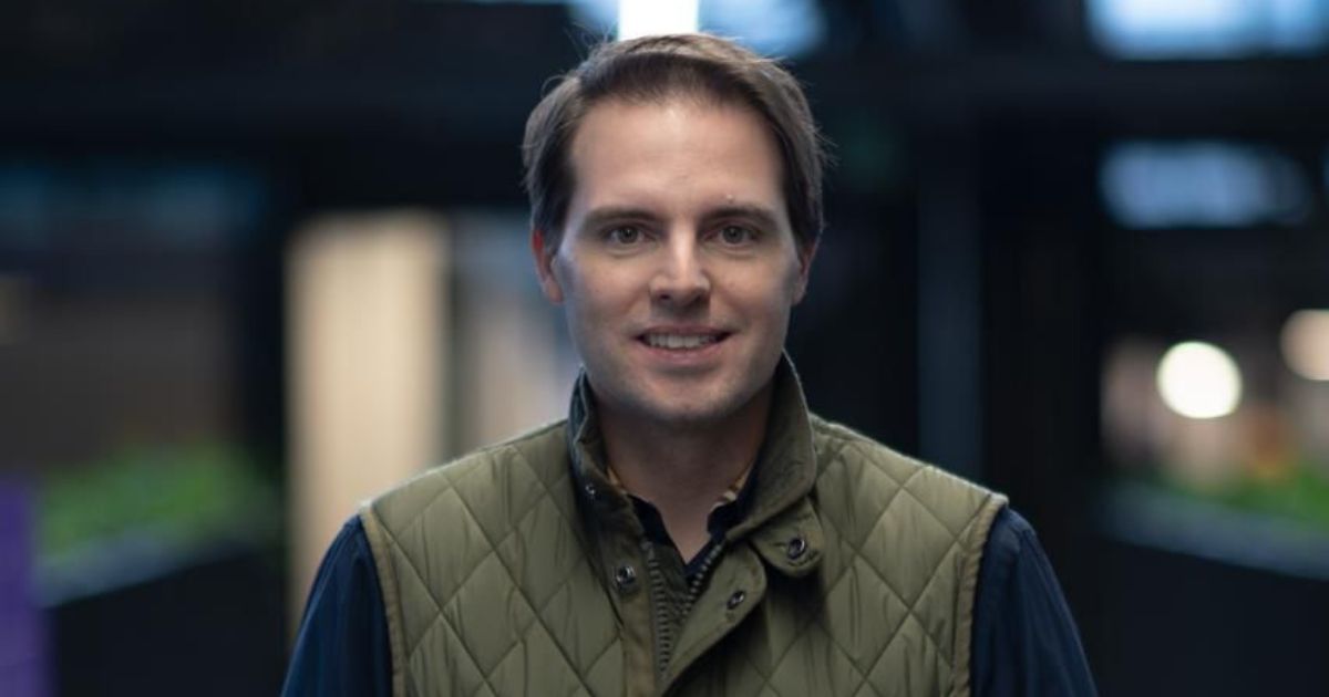 Homem de pele clara e cabelo castanho, levemente penteado para o lado, está sorrindo levemente e olhando para a câmera. Ele veste uma camisa escura com um colete acolchoado verde-oliva. O fundo está desfocado, com iluminação suave e ambiente moderno, sugerindo um espaço interno, como um escritório ou centro de inovação.