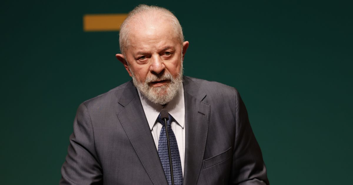 Um homem idoso com barba branca e fato escuro aparece a falar num evento, com expressão séria. O fundo é verde escuro com um elemento gráfico amarelo parcialmente visível. Ele veste camisa branca e gravata azul com padrão.