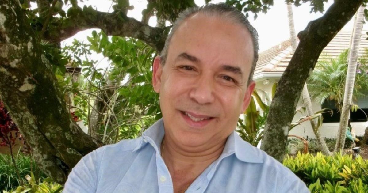 Homem de meia-idade, com cabelo grisalho e camisa azul clara, sorri para a câmara em ambiente exterior. Ao fundo, vê-se um jardim tropical com árvores, plantas e parte de uma casa com telhado inclinado, criando um cenário acolhedor e natural. (Tricentis)