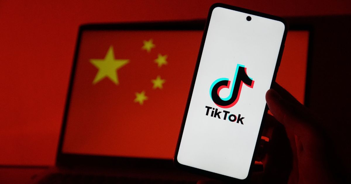 Mão a segurar um smartphone com o logótipo da aplicação TikTok visível no ecrã, sobre fundo branco. Ao fundo, desfocado, vê-se o ecrã de um computador portátil com a bandeira da China, destacando-se o fundo vermelho e cinco estrelas amarelas. A iluminação geral da cena é em tons de vermelho, criando uma ligação visual entre a aplicação e o contexto geopolítico.