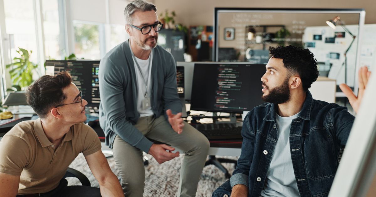 Três homens sentados numa sala de escritório moderna, envolvidos numa conversa técnica. Dois deles são mais jovens e usam roupa casual; o terceiro, com cabelo grisalho e óculos, tem um visual mais formal com camisola e crachá ao pescoço. Ao fundo, dois monitores exibem código de programação. A atmosfera é descontraída e colaborativa, típica de um ambiente de trabalho em tecnologia ou desenvolvimento de software.