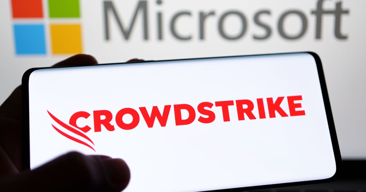 Mão a segurar um smartphone com o logótipo da CrowdStrike em letras vermelhas sobre fundo branco, com o logótipo da Microsoft desfocado ao fundo.