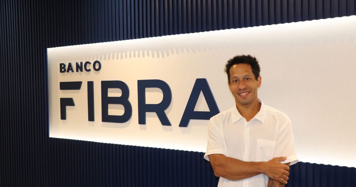 Um homem com camisa branca sorri de braços cruzados em frente a uma parede com o logótipo do "Banco Fibra" em letras grandes. O ambiente é moderno, com iluminação embutida e design de parede com ripas verticais.