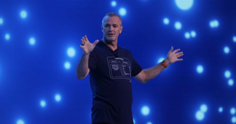 Orador Jeff Clark, COO da Dell, a meio de uma apresentação em palco, com um fundo azul animado por luzes circulares brilhantes. O homem, de meia-idade, está a usar uma t-shirt escura com gráficos e fala com expressividade, gesticulando com os braços abertos.