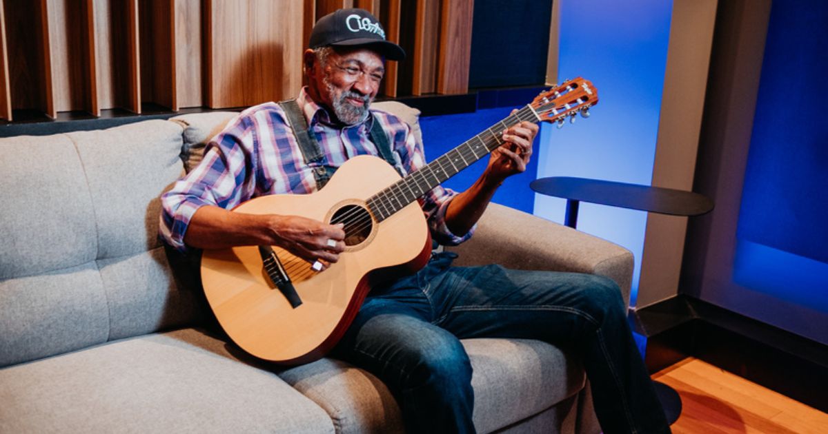 Homem mais velho sentado num sofá cinzento, a tocar um violão acústico com expressão serena e sorridente. Veste uma camisa de flanela às riscas em tons roxo e azul, calças de ganga e boné escuro com a palavra 'Clarks' escrita. O ambiente é moderno e acolhedor, com iluminação azul suave e painéis acústicos na parede, sugerindo que se trata de um estúdio de gravação.