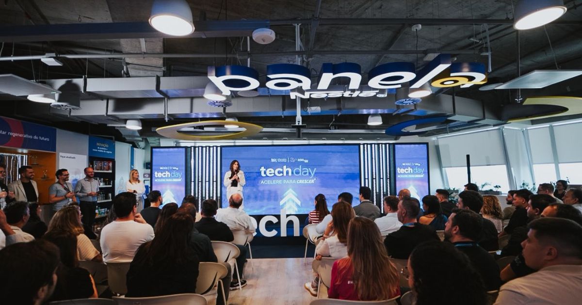 Evento corporativo "Tech Day" realizado no espaço "Panela Nestlé", com uma mulher a discursar no palco diante de um público atento. O ambiente é moderno e bem iluminado, com ecrãs a exibir o tema "Acelere para crescer". O espaço tem decoração tecnológica e branding visível da Nestlé.