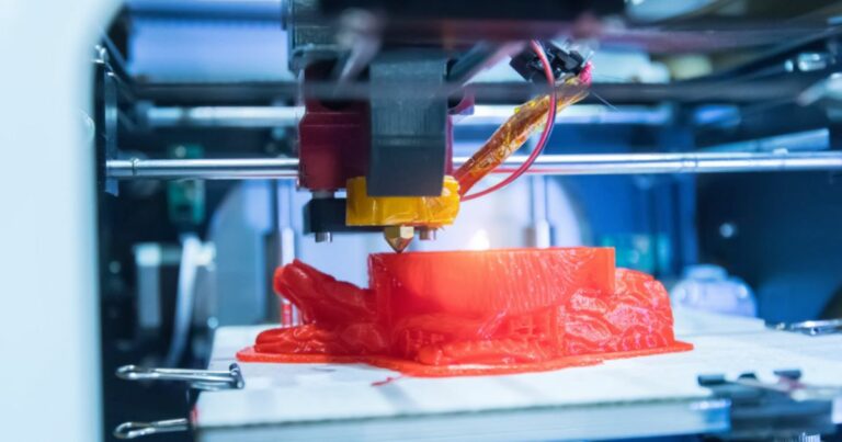 Close-up de uma impressora 3D em funcionamento, a criar um pulmão vermelho em plástico. A cabeça de extrusão está ativa, libertando o material em camadas sobre a plataforma de impressão. A imagem transmite inovação e tecnologia de fabrico digital.