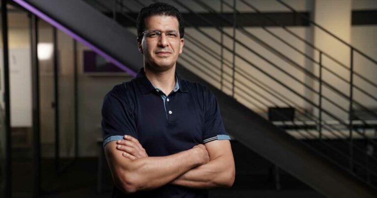 Homem de pele clara, cabelos curtos e escuros, usando óculos e camisa polo azul escuro, está de braços cruzados e olhando para a câmera com expressão confiante. Ele está em um ambiente corporativo moderno, com escada metálica e paredes de vidro ao fundo. A imagem mostra Youseef Lahrech, presidente e COO do Nubank