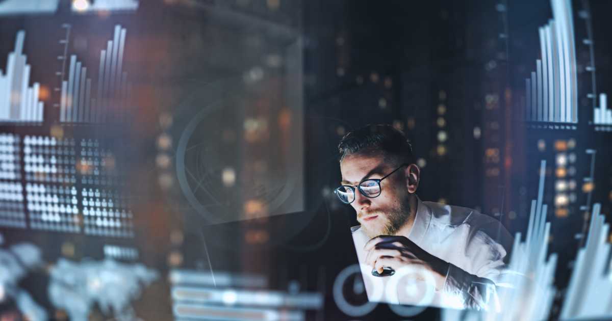 Imagem de um homem usando óculos e camisa clara, concentrado diante de um computador, segurando uma xícara. Ele está cercado por gráficos e elementos visuais digitais projetados, sugerindo análise de dados ou tomada de decisão estratégica em um ambiente de tecnologia. A cena remete ao trabalho de um executivo de tecnologia ou CIO Chief Information Officer