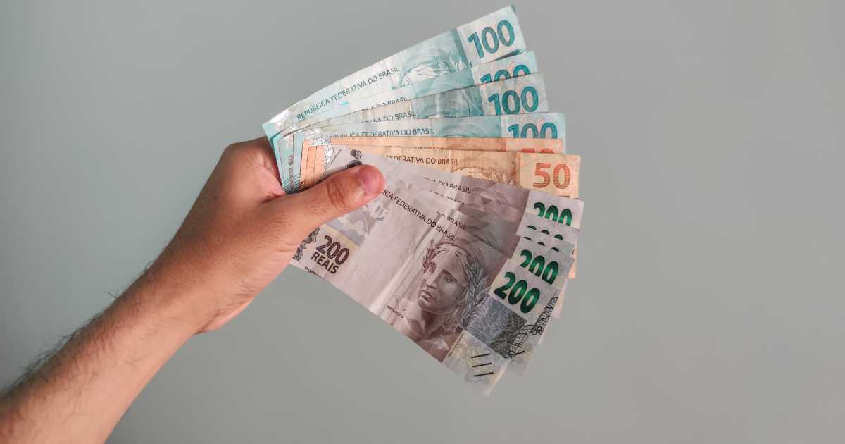 Mão segurando várias cédulas de real brasileiro em leque, com notas de 50, 100 e 200 reais visíveis, sobre fundo cinza neutro. (fintechs, open finance)