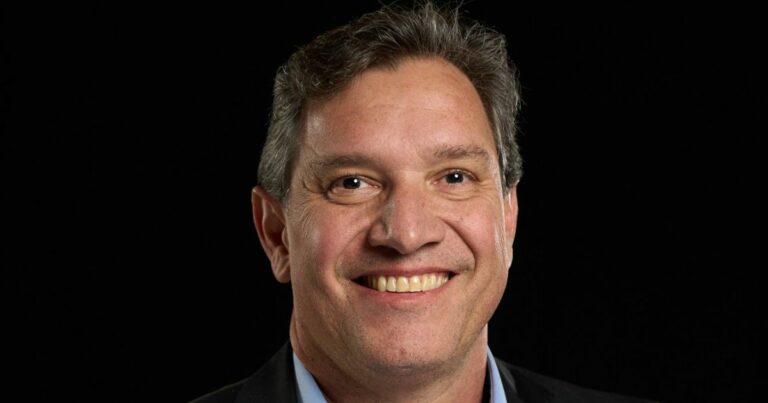 André Facciolli, CEO da Netbr, sorri em retrato com fundo preto, vestindo paletó escuro e camisa azul clara.