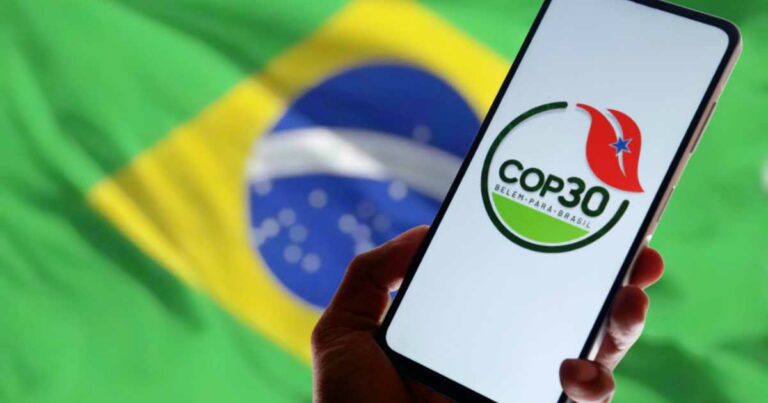 Imagem mostra uma mão segurando um celular que exibe o logotipo oficial da COP30, com os dizeres "COP30 Belém - Pará - Brasil", acompanhado de uma folha estilizada com uma estrela vermelha. Ao fundo, desfocada, está a bandeira do Brasil. A imagem faz referência à realização da 30ª, Conferência das Nações Unidas sobre Mudanças Climáticas (COP30), que ocorrerá em Belém, no estado do Pará, Brasil. Tecnologia