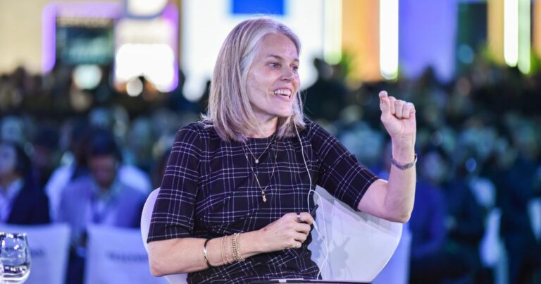 Mulher loira de cabelos lisos e médios sorri enquanto fala em um evento. Ela está sentada, usando um vestido escuro com estampa xadrez branca e vários acessórios dourados (pulseiras, anéis e colares). Ela segura um microfone de lapela preso à roupa e está com o punho direito levemente erguido, em gesto expressivo. Ao fundo, há um público desfocado e luzes coloridas, sugerindo um ambiente de conferência ou painel de discussão. (MIT)