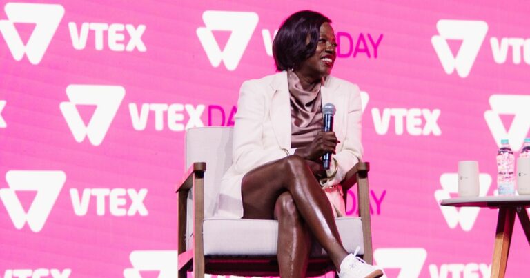 Atriz Viola Davis está sentada em uma cadeira, segurando um microfone. Ao fundo, há um painel rosa com várias repetições do logotipo da VTEX e o texto "VTEX DAY". À direita da pessoa, há uma pequena mesa com duas garrafas e um copo.