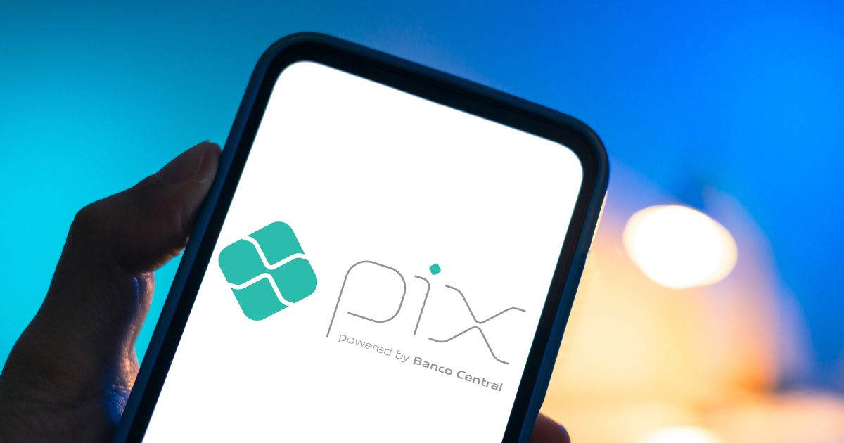 Uma mão segura um smartphone cuja tela exibe o logotipo do Pix, sistema de pagamentos instantâneos do Banco Central do Brasil. O fundo está desfocado, com iluminação em tons de azul e amarelo.