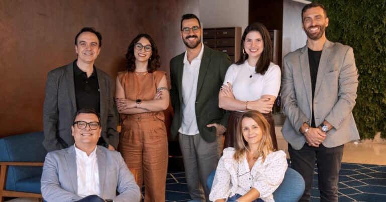 Sete pessoas posam sorrindo para a foto em um ambiente interno elegante e moderno, com decoração em tons terrosos e plantas ao fundo. Quatro delas estão em pé na parte de trás e três estão sentadas à frente. O grupo é composto por homens e mulheres vestindo roupas sociais e casuais-chiques. Duas mulheres usam óculos, uma delas com macacão caramelo. Um homem de blazer verde escuro e camisa branca sorri ao centro, ao lado de uma mulher de blusa branca. À frente, um homem com blazer cinza claro está sentado ao lado de uma mulher de blusa branca com babados. O clima da imagem é descontraído e profissional. (LLYC) (Digital Solvers)