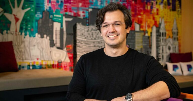 Uma pessoa está sentada com os braços cruzados, vestindo uma camisa preta de manga longa e um relógio no pulso esquerdo. O rosto da pessoa está desfocado. Ao fundo, há um mural colorido com diversos elementos abstratos, incluindo silhuetas de pessoas, prédios e cores vibrantes. (PayPal)