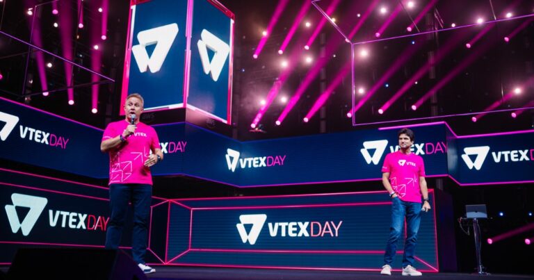 Duas pessoas estão em um palco iluminado por luzes rosa e roxas, com várias telas ao fundo exibindo o texto "VTEX DAY" e o logotipo da VTEX. Ambas vestem camisetas rosa com o logotipo da VTEX e padrões geométricos. O ambiente transmite uma atmosfera vibrante e tecnológica, típica de eventos corporativos ou conferências de inovação.
