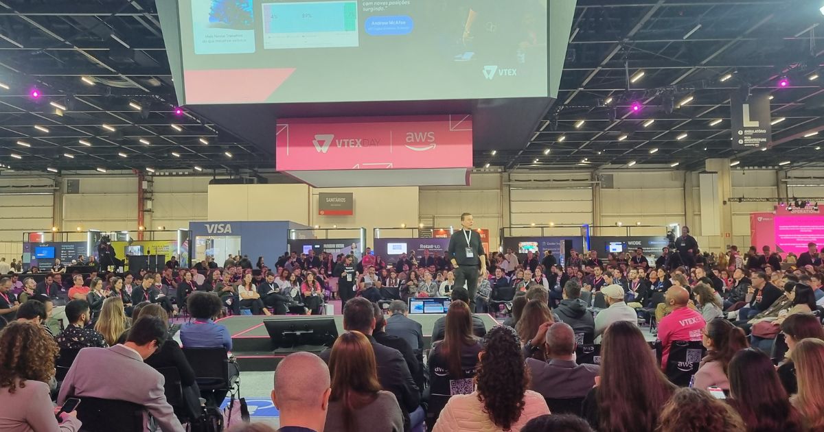 A imagem retrata um grande evento corporativo ou conferência, com um público numeroso sentado e voltado para um palco principal. No palco, um palestrante apresenta diante de uma tela ampla que exibe slides com os logotipos da VTEX e da AWS. O ambiente é amplo, com pé-direito alto e iluminação profissional. Ao fundo, é possível ver estandes de empresas, incluindo um com o logotipo da VISA. Os participantes parecem atentos, alguns fazendo anotações ou utilizando dispositivos eletrônicos. (Google)