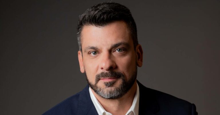 Homem de cabelo e barba curtos e escuros, usando paletó azul-marinho e camisa branca, posando em frente a um fundo neutro. Na imagem está Renato Simões, diretor de Produtos e Marketing da Oi Soluções.