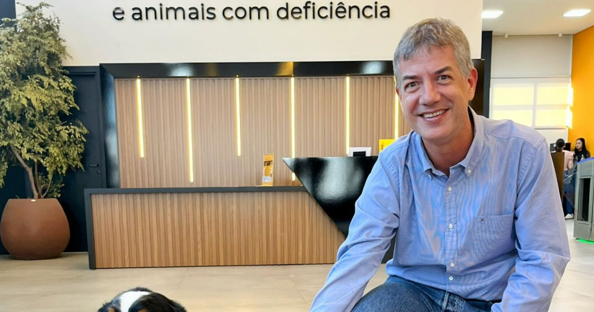 Fernando Rostock, novo head de TI da Adimax, aparece sorridente em uma área interna moderna da empresa, vestindo camisa azul clara e jeans. Ele está agachado ao lado de um cachorro, parcialmente visível no canto inferior esquerdo da imagem. Ao fundo, há um balcão de recepção com detalhes em madeira e uma parede com a frase “e animais com deficiência”, além de uma planta decorativa em vaso grande