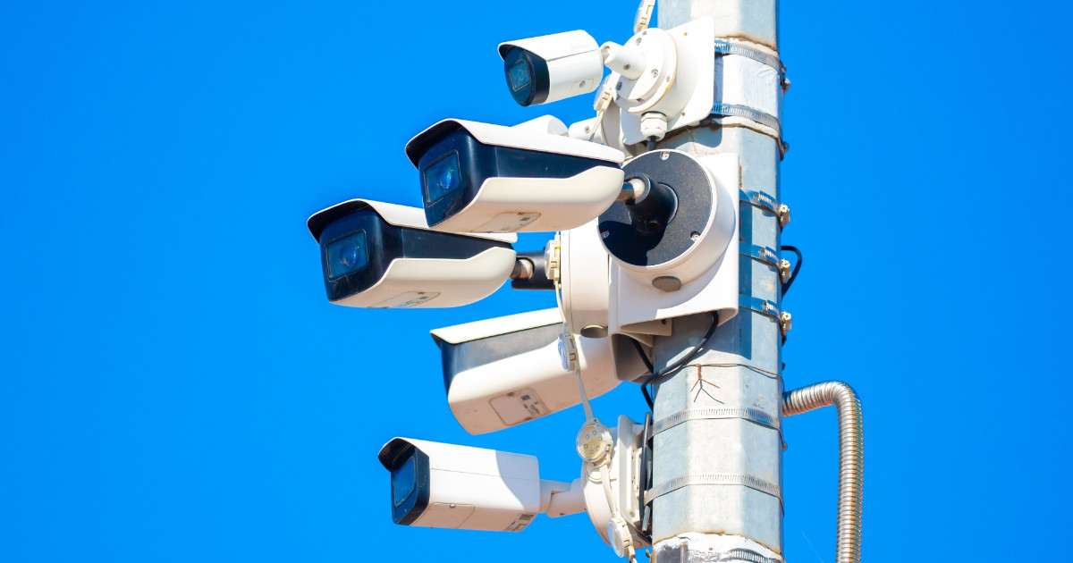 Conjunto de câmeras de vigilância instaladas em um poste metálico, apontadas em diferentes direções, sob um céu azul claro. A imagem representa um sistema de monitoramento urbano ou controle de tráfego com tecnologia de segurança eletrônica (Foucault)