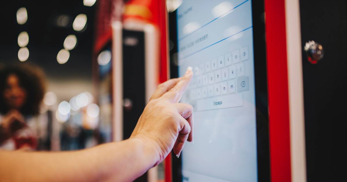 Pessoa interage com um totem eletrônico do iFood, com a interface da empresa Videosoft visível na tela. A mão está digitando em um teclado virtual exibido no display sensível ao toque. O ambiente ao fundo está desfocado, sugerindo um local público movimentado, como um shopping ou praça de alimentação