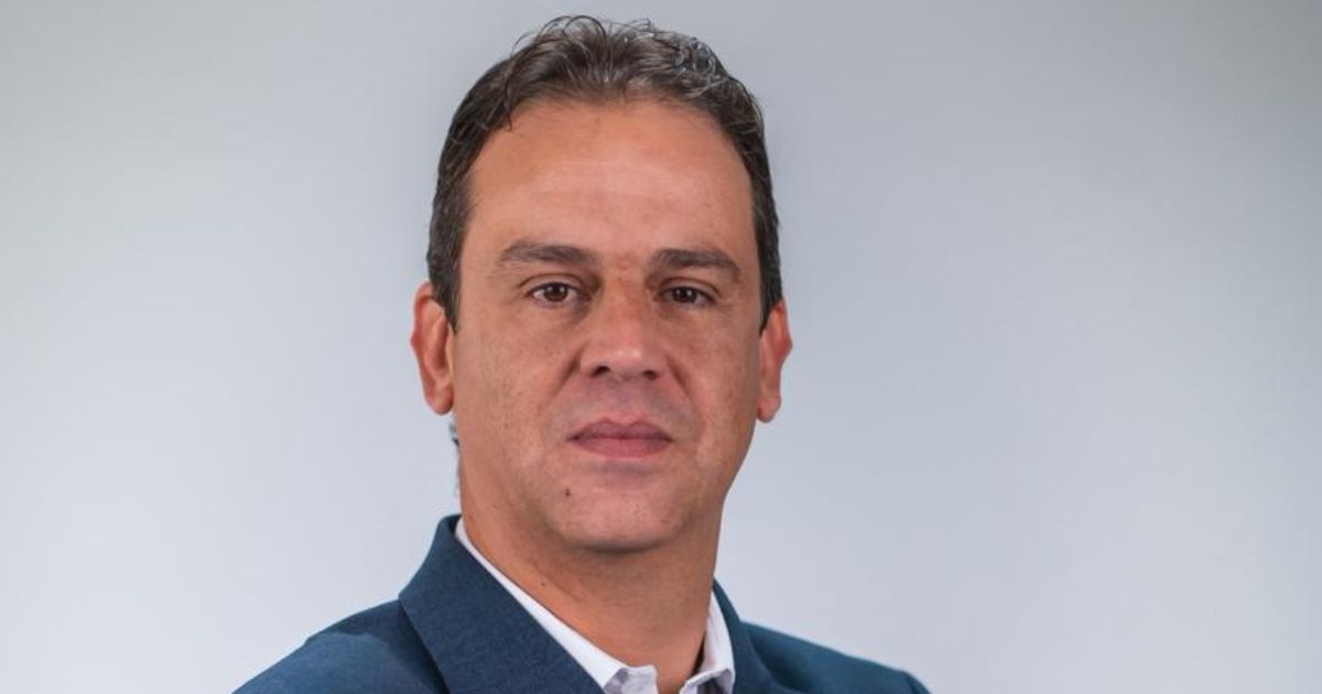 Pessoa em ambiente formal, vestindo paletó azul escuro sobre camisa branca. O fundo é claro e simples, destacando a figura principal. A imagem pode ser usada em contextos profissionais ou institucionais, como identificação ou apresentação. (RH)