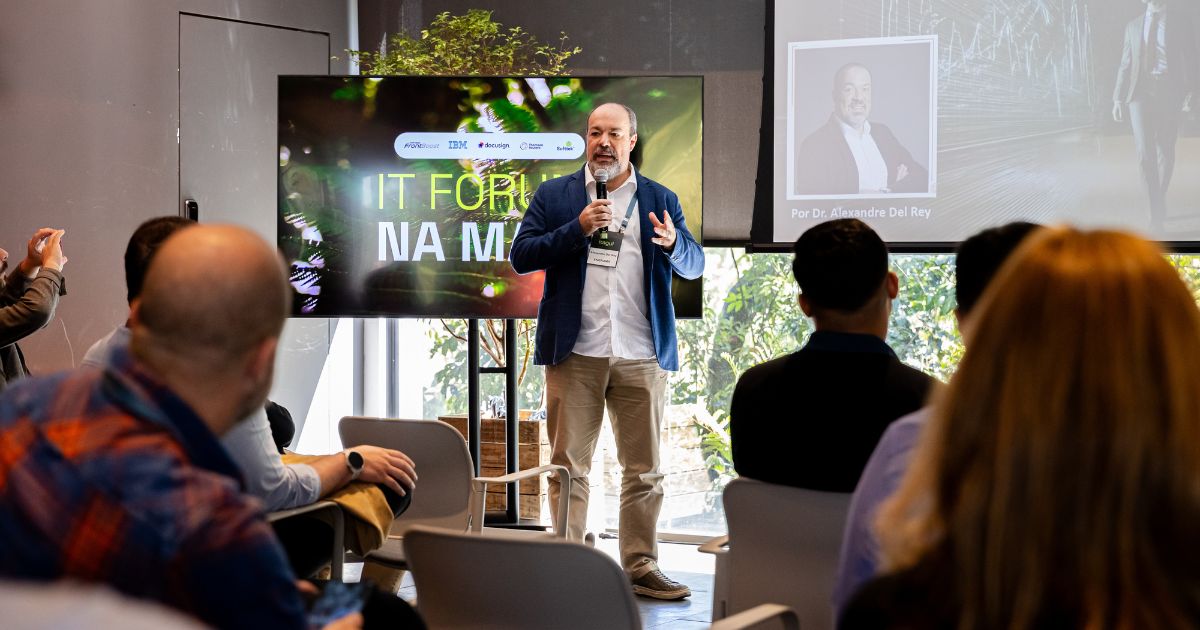 A foto mostra uma pessoa em pé, segurando um microfone e falando para um grupo de pessoas sentadas em cadeiras. A pessoa veste um blazer azul, camisa branca e calça clara, com um crachá pendurado no pescoço. Ao fundo, há uma tela grande exibindo uma imagem com folhas verdes e texto parcialmente visível, incluindo “IT FÓRUM NA MA...”. Acima do texto, aparecem logotipos de empresas. À direita, há outra tela projetando uma imagem com gráficos e uma foto de outra pessoa, acompanhada do texto “Por Dr. Alexandre Del Rey”. O ambiente parece ser uma sala de conferências iluminada por luz natural, com janelas mostrando vegetação externa. (IA)