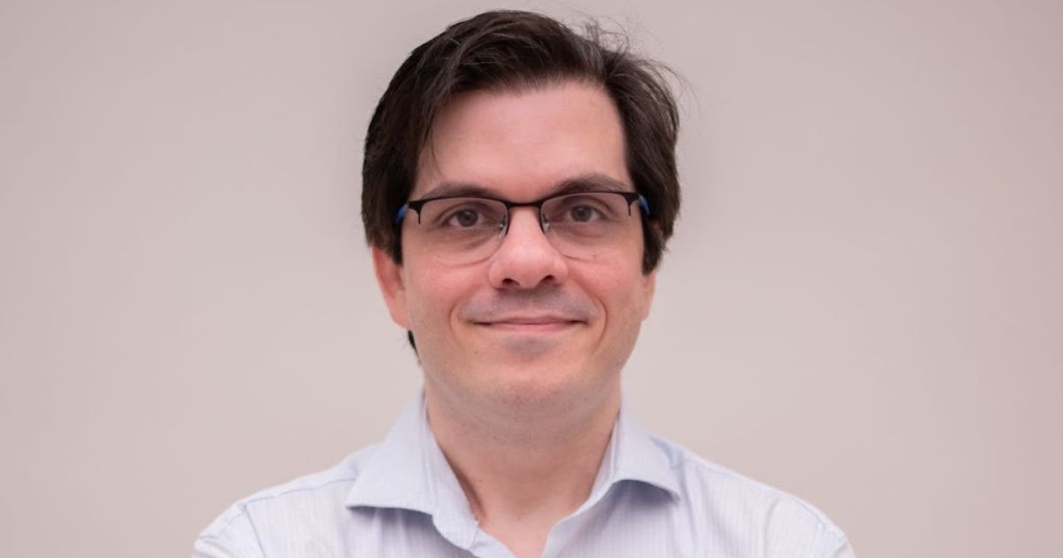 A imagem mostra uma pessoa vestindo uma camisa social de cor clara, com colarinho e botões visíveis. O cabelo é escuro e está penteado para trás, com um aspecto levemente volumoso. O fundo é simples e uniforme, em tom claro, sugerindo um ambiente neutro ou estúdio fotográfico. A pessoa está posicionada de frente, com os braços cruzados à altura do peito. ( Flutter Brazil )