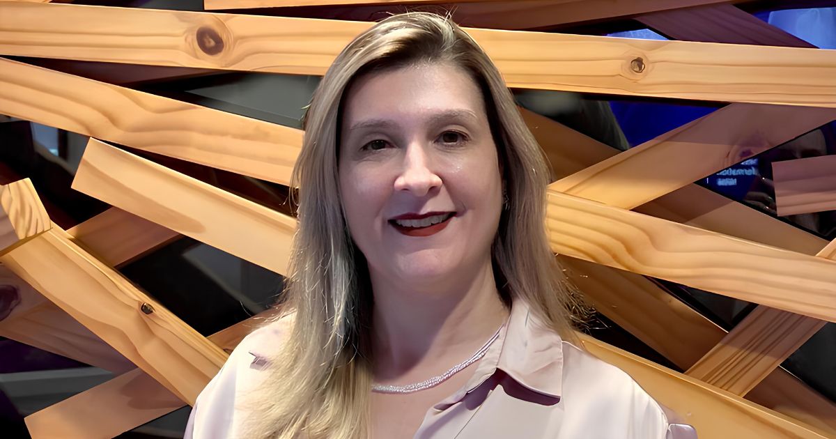 A imagem mostra uma pessoa vestindo uma camisa de tecido acetinado em tom claro, com gola tradicional e botões parcialmente visíveis. Ao redor do pescoço, há um colar delicado, composto por pequenas contas ou pedras claras. O fundo é formado por uma estrutura artística de ripas de madeira dispostas em ângulos cruzados, criando um padrão geométrico interessante. Entre as ripas, é possível ver áreas escuras e reflexos que sugerem um ambiente interno com iluminação controlada. (transforma)