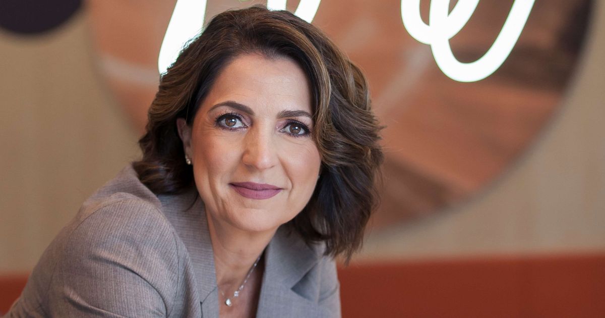 Pessoa com cabelo curto e ondulado, vestindo blazer cinza, olha diretamente para a câmera. Ao fundo, há um objeto circular desfocado com texto cursivo branco parcialmente legível. A imagem apresenta um estilo de retrato profissional, com foco na pessoa em primeiro plano. (AWS)