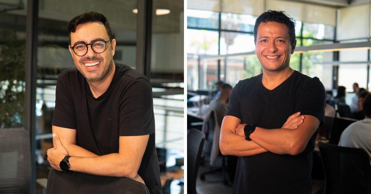 A imagem mostra duas pessoas lado a lado em um ambiente de escritório moderno. Ambas estão usando camisetas pretas e óculos, e posam sorrindo com os braços cruzados. Ao fundo, é possível ver paredes de vidro, mesas e outras pessoas trabalhando, sugerindo um espaço corporativo colaborativo. (fintech)