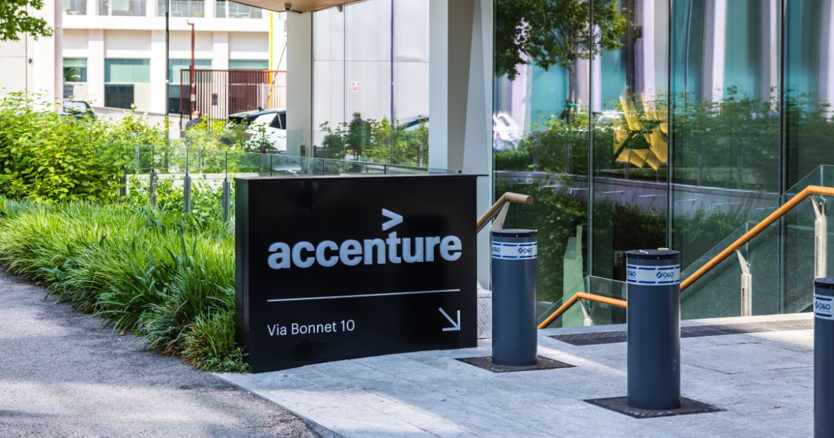 A imagem mostra a fachada de um prédio da empresa Accenture. Em destaque, há uma placa preta com o logotipo da Accenture — incluindo uma seta voltada para a direita — e o endereço "Via Bonnet 10". A placa está posicionada ao lado de um caminho que leva à entrada do edifício, que possui paredes de vidro e vegetação ao lado. Na frente da entrada, há três barreiras cilíndricas com rótulos ou anúncios visíveis.
