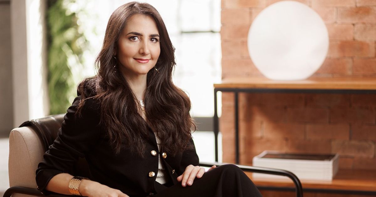 A imagem mostra uma pessoa sentada em uma cadeira em um ambiente interno. Ela está vestindo uma roupa escura com botões dourados e acessórios. Ao fundo, há uma parede de tijolos, uma estante com livros e um objeto redondo branco. A composição transmite uma atmosfera profissional e organizada. (Anima)