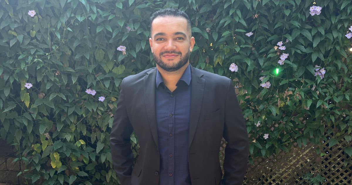 A imagem mostra uma pessoa em pé diante de um fundo verde vibrante, composto por folhas e pequenas flores roxas. Ela veste um blazer escuro sobre uma camisa também escura e está voltada diretamente para a câmera. O cenário parece ser ao ar livre, como um jardim ou parque, oferecendo um contraste natural à apresentação formal da pessoa. (startup, IA)