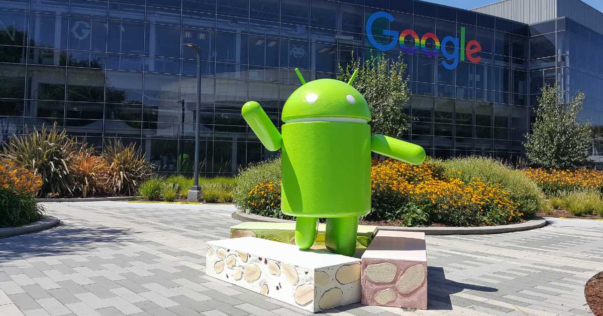 Estátua grande do mascote verde do Android em área externa ajardinada, posicionada sobre base decorada, com o prédio envidraçado do Google ao fundo e jardins com flores amarelas ao redor
