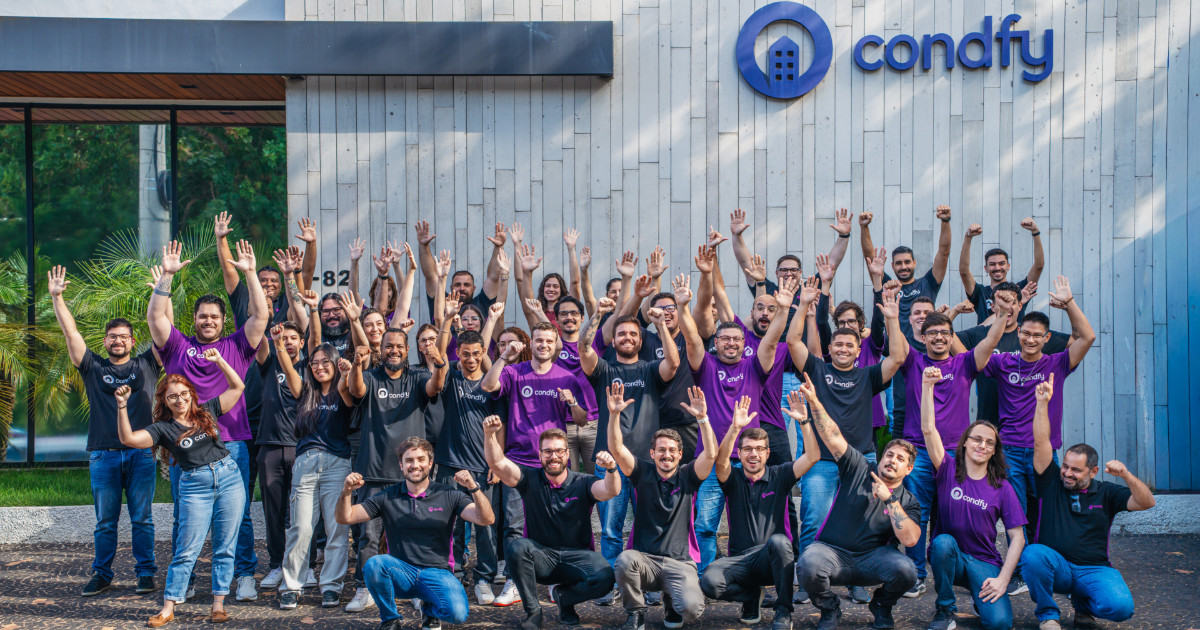 **Texto alternativo sugerido:** Foto de um grande grupo de colaboradores da Condfy posando em frente ao prédio da empresa, todos sorrindo e comemorando com braços levantados. Eles vestem camisetas pretas e roxas com o logo da Condfy, formando uma imagem vibrante de equipe unida diante da fachada moderna da empresa.