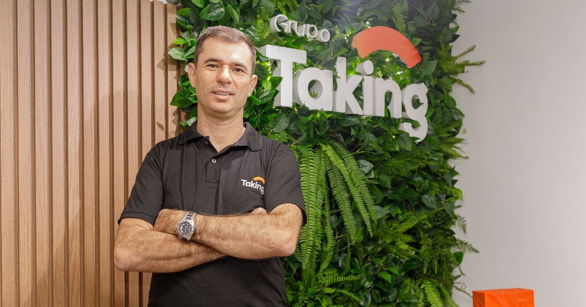 Marco Romero, CEO do Grupo Taking, aparece em pé, de braços cruzados, usando uma camisa polo preta com o logo da empresa. Ele está diante de uma parede verde composta por plantas naturais e o logotipo do Grupo Taking em branco com um arco laranja. A imagem é bem iluminada e tem caráter institucional. Foto: Divulgação.