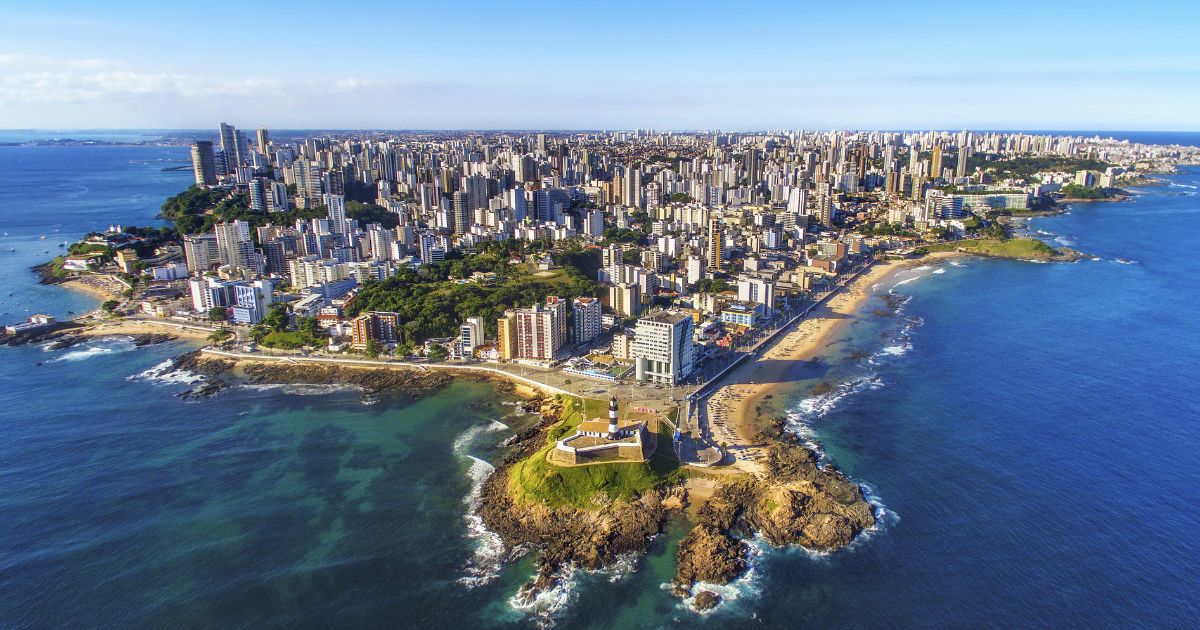 A foto mostra uma vista aérea de uma cidade litorânea com uma extensa faixa de prédios altos ao fundo e várias praias ao longo da costa. Em primeiro plano, há uma pequena península com um farol histórico e construções ao redor, cercada por águas azuis e áreas rochosas. O mar é amplo e ocupa grande parte da imagem, com tons que variam do verde próximo à costa ao azul profundo mais distante. O céu está claro, indicando um dia ensolarado. (futuro)