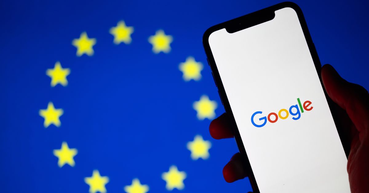 A foto mostra um smartphone sendo segurado, exibindo na tela o logotipo do Google em letras coloridas sobre fundo branco. Ao fundo, desfocado, há a bandeira da União Europeia, composta por um círculo de estrelas amarelas sobre fundo azul. A imagem sugere um contexto relacionado à tecnologia, privacidade ou regulamentações digitais.
