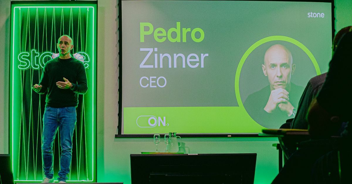 A imagem mostra um evento corporativo com identidade visual predominante em tons de verde, associado à marca Stone. No palco, há uma pessoa apresentando, posicionada à esquerda, diante de um painel iluminado com linhas verticais e o logotipo da empresa. À direita, um grande telão exibe informações do palestrante: Texto visível: “Pedro Zinner” em destaque, com “CEO” logo abaixo. Logotipo da Stone no canto superior direito. Elemento gráfico circular com a foto do palestrante. Palavra “ON.” na parte inferior do slide. No primeiro plano, há uma mesa com copos e garrafas de água, sugerindo um ambiente preparado para uma apresentação formal. A iluminação é intensa, com foco no verde, reforçando a identidade da marca e criando um clima moderno e tecnológico.