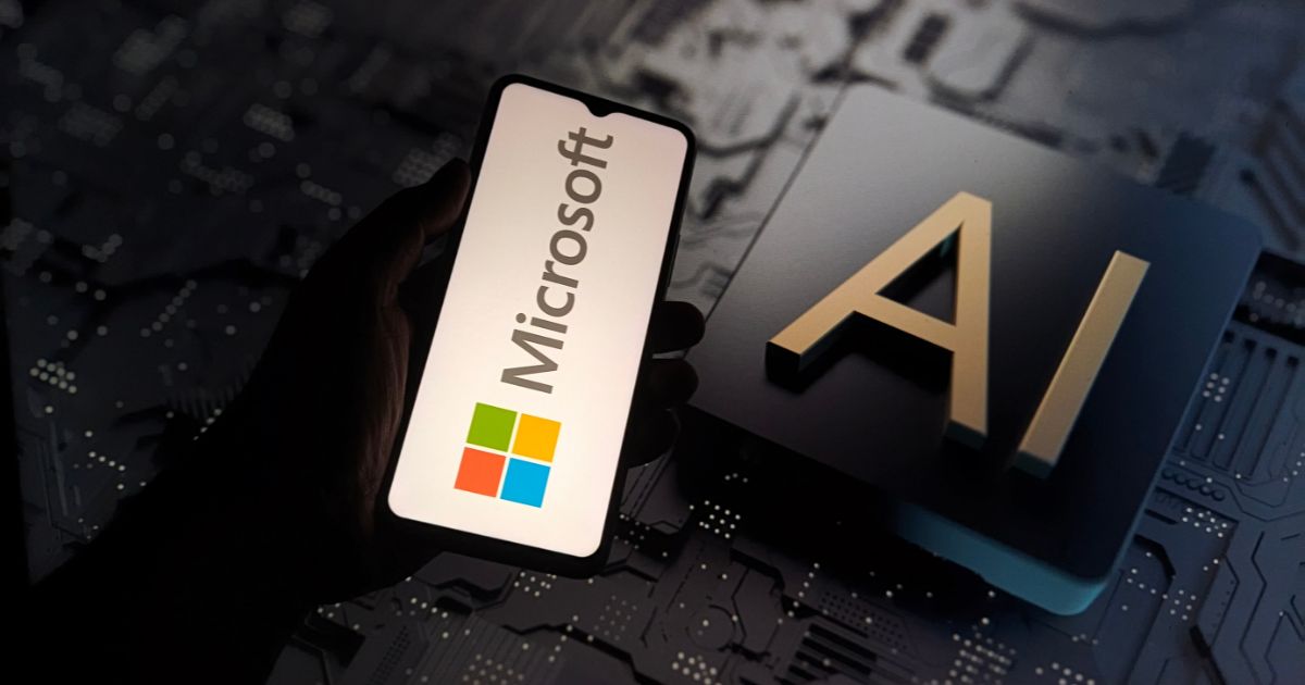 A cena mostra um smartphone sendo segurado por uma mão, exibindo a tela com o logotipo da Microsoft e seu ícone colorido característico. Ao lado, sobre uma superfície com padrão de circuito eletrônico, há uma peça tridimensional com as letras “AI” em destaque, indicando inteligência artificial. O fundo é escuro, com iluminação suave que realça os elementos tecnológicos, transmitindo uma atmosfera moderna e voltada para inovação digital. (superinteligência)