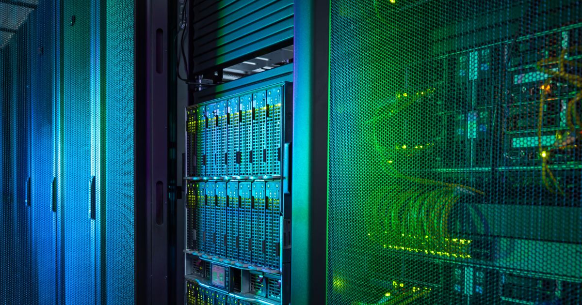 A foto mostra o interior de um data center com várias racks de servidores. À esquerda, há gabinetes fechados com iluminação azul, enquanto à direita aparecem servidores com painéis metálicos perfurados e cabos organizados, iluminados por luz verde. A imagem destaca a infraestrutura tecnológica, com componentes eletrônicos e sistemas de refrigeração visíveis, transmitindo um ambiente moderno e de alta capacidade computacional. (startup, tiktok)