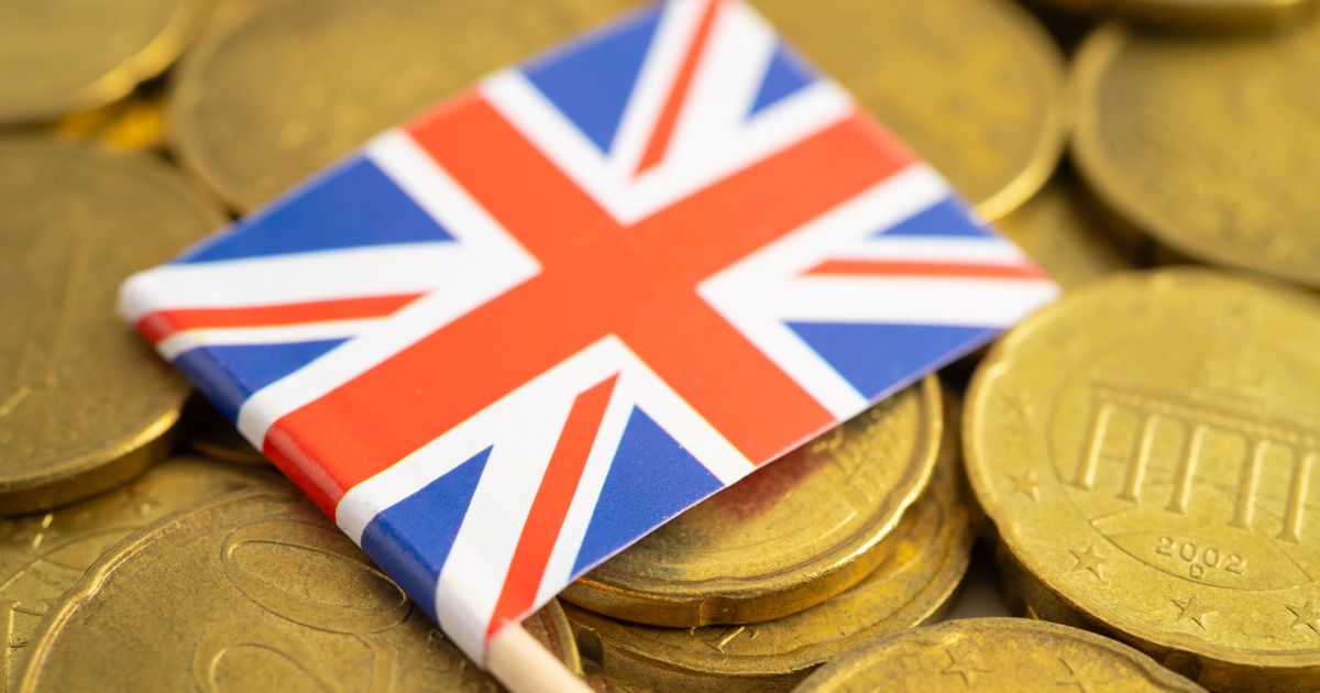A foto mostra uma pequena bandeira do Reino Unido (Union Jack) apoiada sobre várias moedas douradas empilhadas. As moedas têm detalhes gravados, incluindo símbolos arquitetônicos e números, sugerindo que sejam moedas europeias. O fundo é preenchido pelas moedas, criando um contraste entre o dourado e as cores vibrantes da bandeira (vermelho, azul e branco).